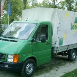 Zielony bus Volkswagen LT z plandeką Landi na parkingu. Widoczne logo marki na plandece i przednia część pojazdu. Tło stanowią drzewa i ogrodzenie.
