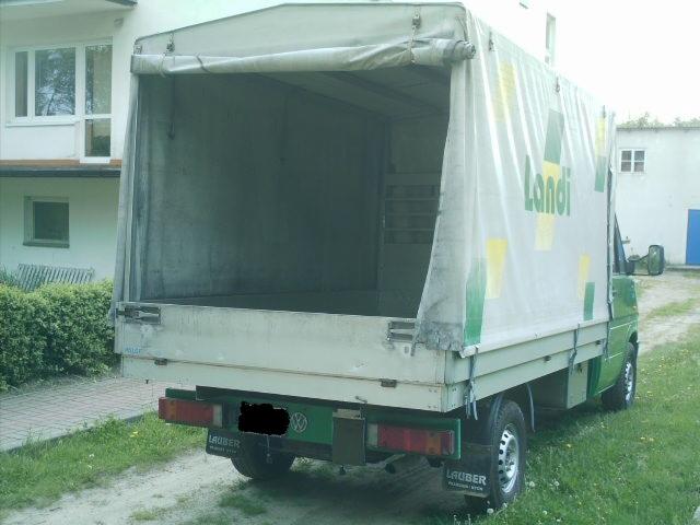 Widok z tyłu zielono-białego busa dostawczego z otwartą przestrzenią ładunkową i plandeką z logo Landi, zaparkowanego na trawie przed budynkiem.