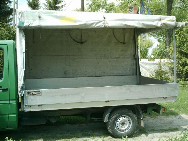 Zielony bus z otwartą skrzynią ładunkową i plandeką, zaparkowany na trawie w słoneczny dzień.