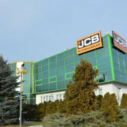 Budynek biurowy z zielonymi panelami i logo JCB Interhandler na dachu, otoczony zielenią i lampą uliczną.