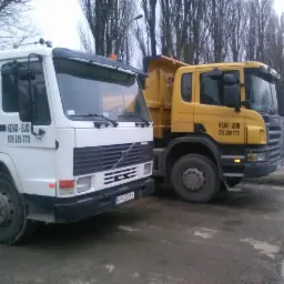 Dwa samochody ciężarowe typu wywrotka, biały Volvo i ż&oacute;łty Scania, z widocznym logo firmy 'KEMO-BUD' i numerem telefonu, zaparkowane na placu budowy w pochmurny dzień.