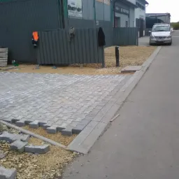 Układanie szarej kostki brukowej typu 'behaton' na podsypce piaskowo-żwirowej, widoczne niezakończone prace, krawężnik oraz samochód w tle.