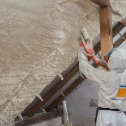 Pracownik w kombinezonie ochronnym aplikuje piankę izolacyjną na poddaszu, widoczne krokwie i warstwa pianki z charakterystycznymi wypustkami, w tle białe bloczki betonowe i logo 'Home Zone 2018'.