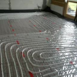 Rozłożone rury ogrzewania podłogowego na folii z siatką, przymocowane plastikowymi klipsami, widok z góry na surowy stan wykończenia wnętrza.