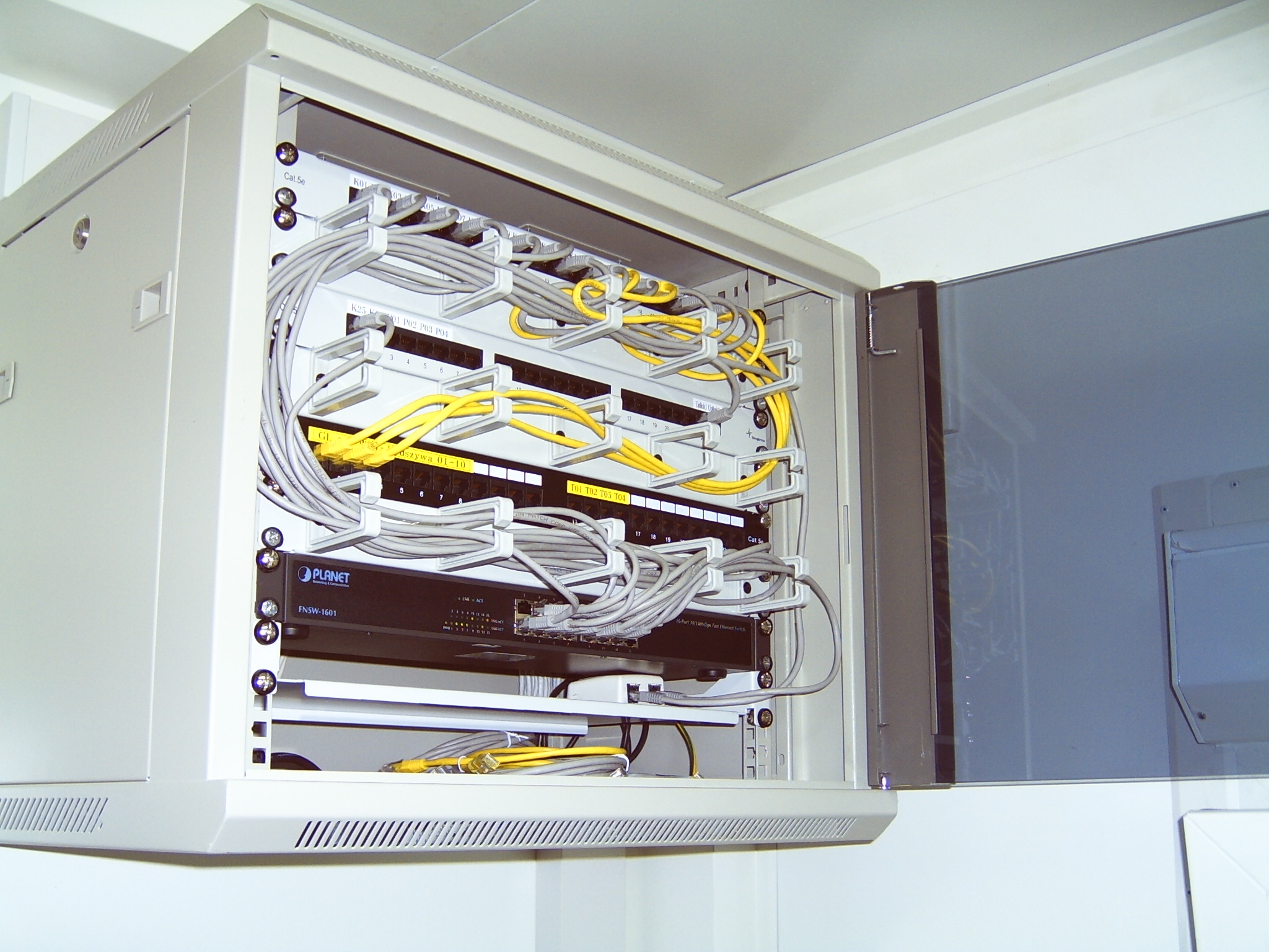 Otwarta szafa serwerowa z zamontowanym switchem Planet FNSW-1601, patch panelem i uporządkowanymi kablami sieciowymi w kolorach szarym i żółtym.
