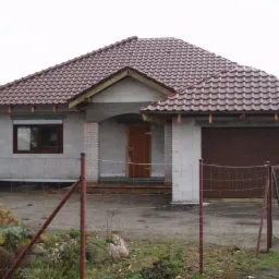 Niedokończony dom z czerwonym dachem z cegły, widoczne bloczki Ytong, brązowa brama garażowa, ogrodzenie z siatki i fragment niebieskiego auta.