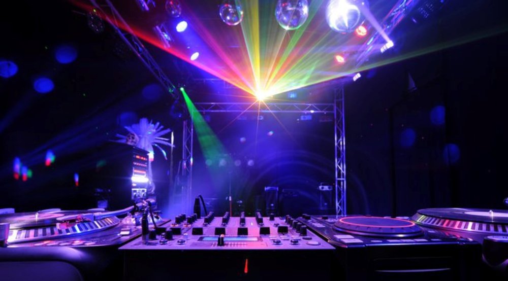 Konsoleta DJ-a z gramofonami i mikserem oświetlona kolorowymi laserami i dyskotekowymi kulami, widok z niskiej perspektywy.