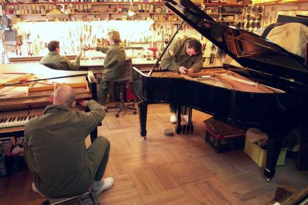 Warsztat renowacji pianin: trzech mężczyzn w kombinezonach naprawia fortepian i pianino, na ścianie w tle wiszą narzędzia.