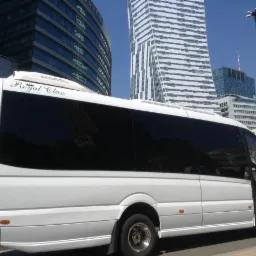 Biały bus 'Royal Class' z otwartymi drzwiami na tle nowoczesnych biurowców Warszawy. Pojazd gotowy do drogi, widoczne detale: logo i dane kontaktowe.