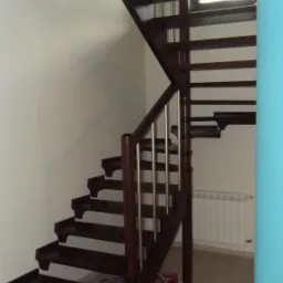 Ciemne drewniane schody z metalowymi poręczami w jasnym wnętrzu, widoczne zabawki pod schodami.