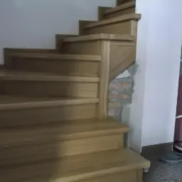 Schody drewniane z jasnego drewna, widoczne niedokończone wykończenie ściany z cegłą i grzejnik z butami w tle.