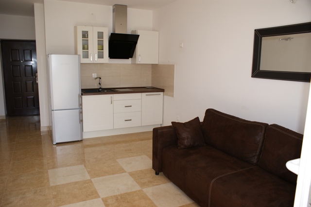 Jasne wnętrze apartamentu z brązową sofą, białą kuchnią i błyszczącą podłogą w szachownicę.