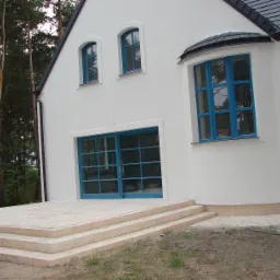 Biały dom z niebieską stolarką okienną i drzwiową, widoczne trzy stopnie prowadzące do tarasu z jasnych płyt, w tle las iglasty.