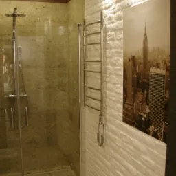 Nowoczesna łazienka z kabiną prysznicową wykończoną kamieniem, grzejnikiem drabinkowym na ścianie z dekoracyjną cegłą i obrazem panoramy miasta.