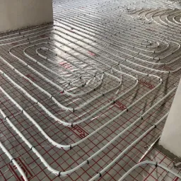 Białe rury ogrzewania podłogowego rozłożone na srebrnej folii z czerwoną siatką, widoczne czarne uchwyty mocujące, surowy stan wykończenia wnętrza.