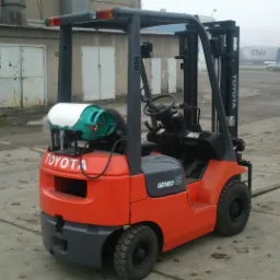 Czerwono-szary wózek widłowy spalinowy Toyota 7FGF15 z butlą gazową z tyłu, stojący na betonowym placu w pochmurny dzień, w tle budynki magazynowe i zaparkowane samochody.