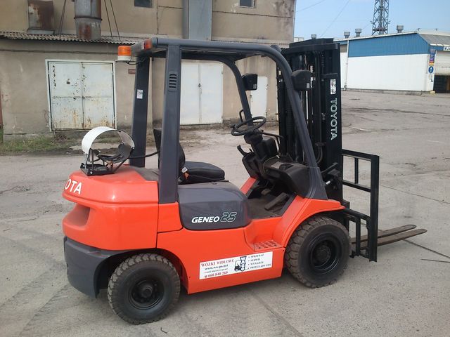Pomarańczowy wózek widłowy Toyota 7FGF25 Geneo 25 z czarnymi oponami i białym kaskiem na tylnej obudowie, stojący na betonowym placu przed budynkiem magazynowym.