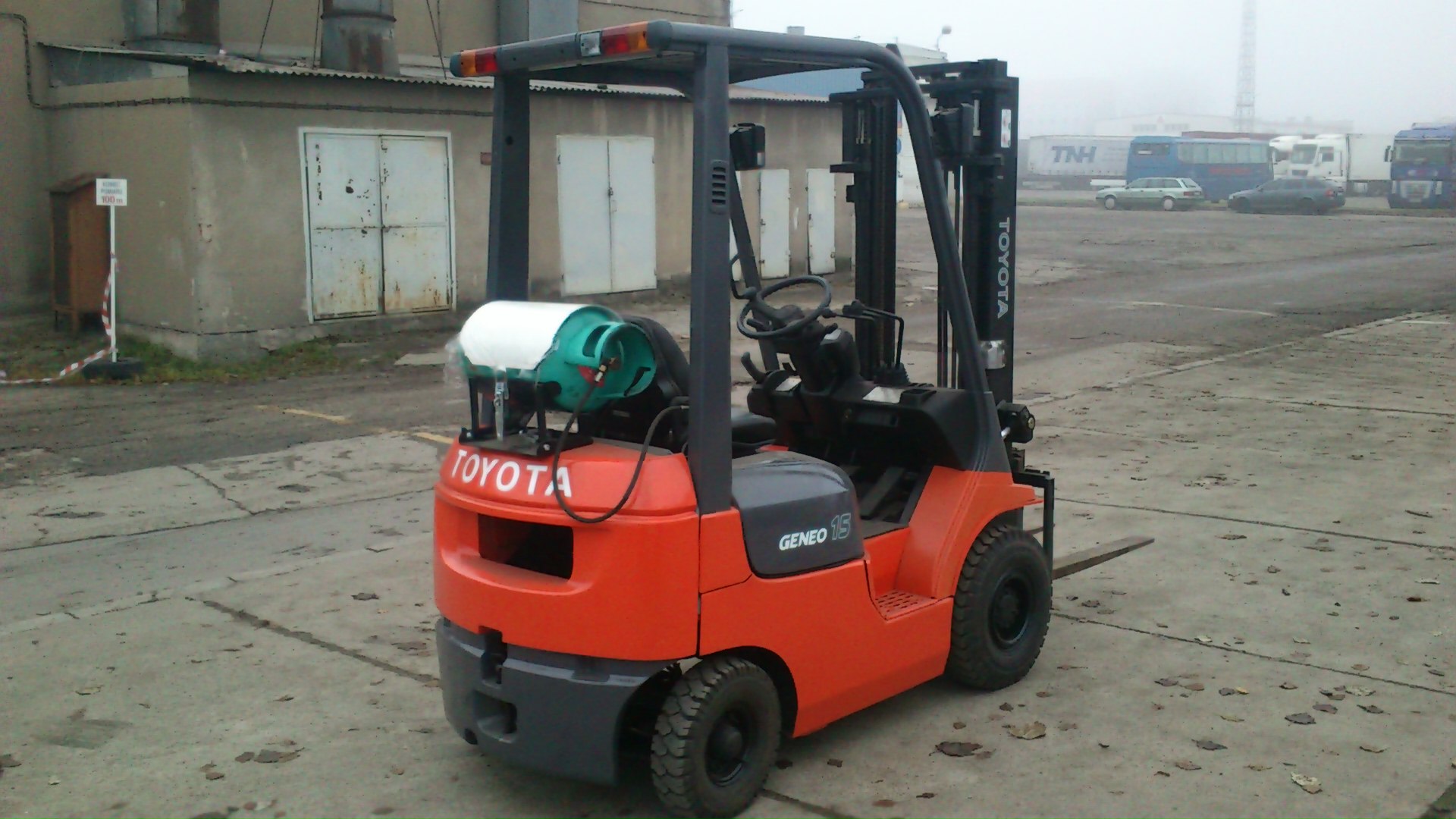 Czerwono-szary wózek widłowy spalinowy Toyota 7FGF15 z butlą gazową z tyłu, stojący na betonowym placu w pochmurny dzień, w tle budynki magazynowe i zaparkowane samochody.