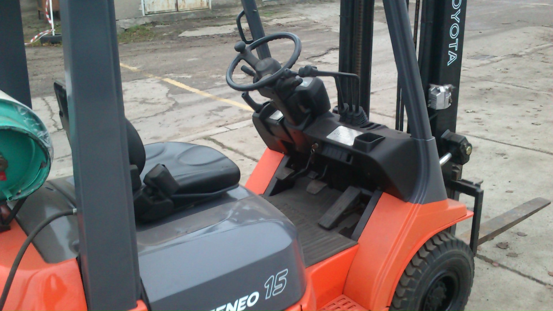 Pomarańczowo-szary wózek widłowy Toyota 7FGF15 z butlą gazową, widoczny kokpit, kierownica i pedały, na zewnątrz.