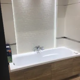 Nowoczesna łazienka: biała wanna z obudową imitującą drewno, chromowana armatura i ściana wyłożona białymi heksagonalnymi płytkami z subtelnym oświetleniem LED.