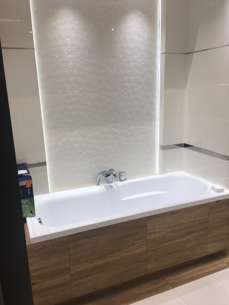 Nowoczesna łazienka: biała wanna z obudową imitującą drewno, chromowana armatura i ściana wyłożona białymi heksagonalnymi płytkami z subtelnym oświetleniem LED.