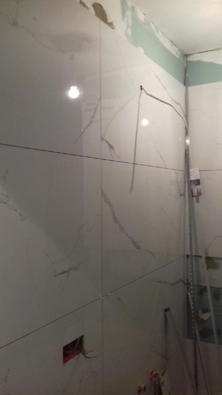 Świeżo położone, duże, białe płytki imitujące marmur na ścianach łazienki w trakcie remontu, widoczne niedoskonałości na suficie, otwór na gniazdko elektryczne i listwa przypodłogowa oparta o ścianę.