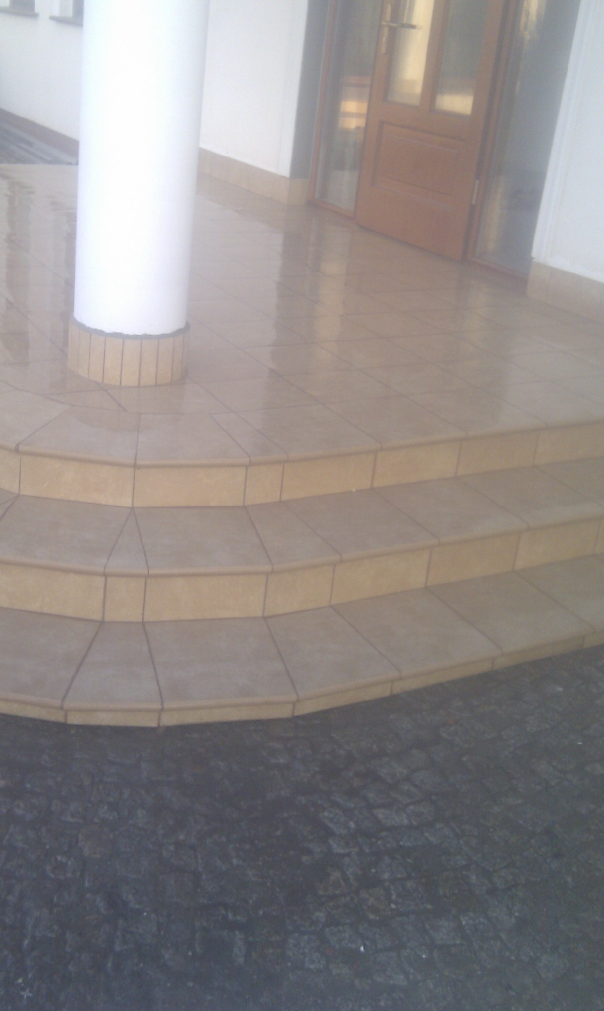 Beżowe płytki na schodach wejściowych z zaokrąglonymi krawędziami, biała kolumna z dekoracyjnym wykończeniem u podstawy, widoczne drzwi wejściowe i fragment chodnika z kostki brukowej.