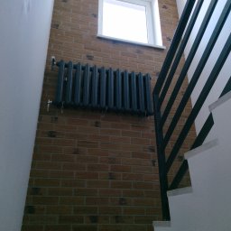 Zakład Ogólnobudowlany - Ceglana ściana ze stalowym grzejnikiem i oknem, widok z dołu. Fragment schodów z czarną balustradą po prawej. Oświetlenie sufitowe widoczne w górnej części kadru.