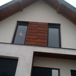 Zakład Ogólnobudowlany - Elewacja domu z oknami i drewnianą okładziną. Widoczne czarne bramy garażowe i drewniane wykończenie dachu. W oknie naklejka.