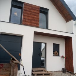 Zakład Ogólnobudowlany - Nowoczesny dom z białą elewacją, drewnianymi akcentami i ciemnymi oknami. Widoczny fragment garażu, wejście i dekoracje okienne. Uwagę przykuwa świeżo posadzona róża.