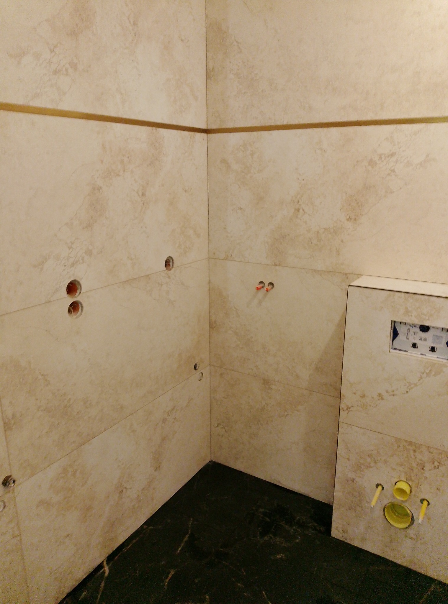 Wykończona ściana łazienki z beżowych płytek z delikatnym wzorem, ozdobiona złotym paskiem. Widoczne otwory na armaturę i fragment podłogi z ciemnego marmuru.