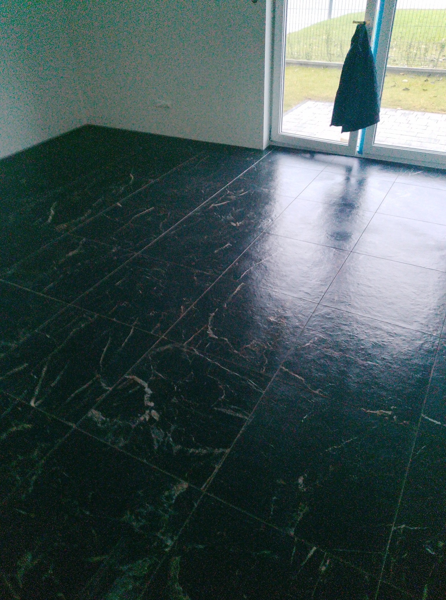 Położona, ciemna, marmurkowa podłoga z płytek w pokoju z oknem. Na oknie wisi ciemna kurtka. Widoczne odbicie światła na powierzchni płytek.