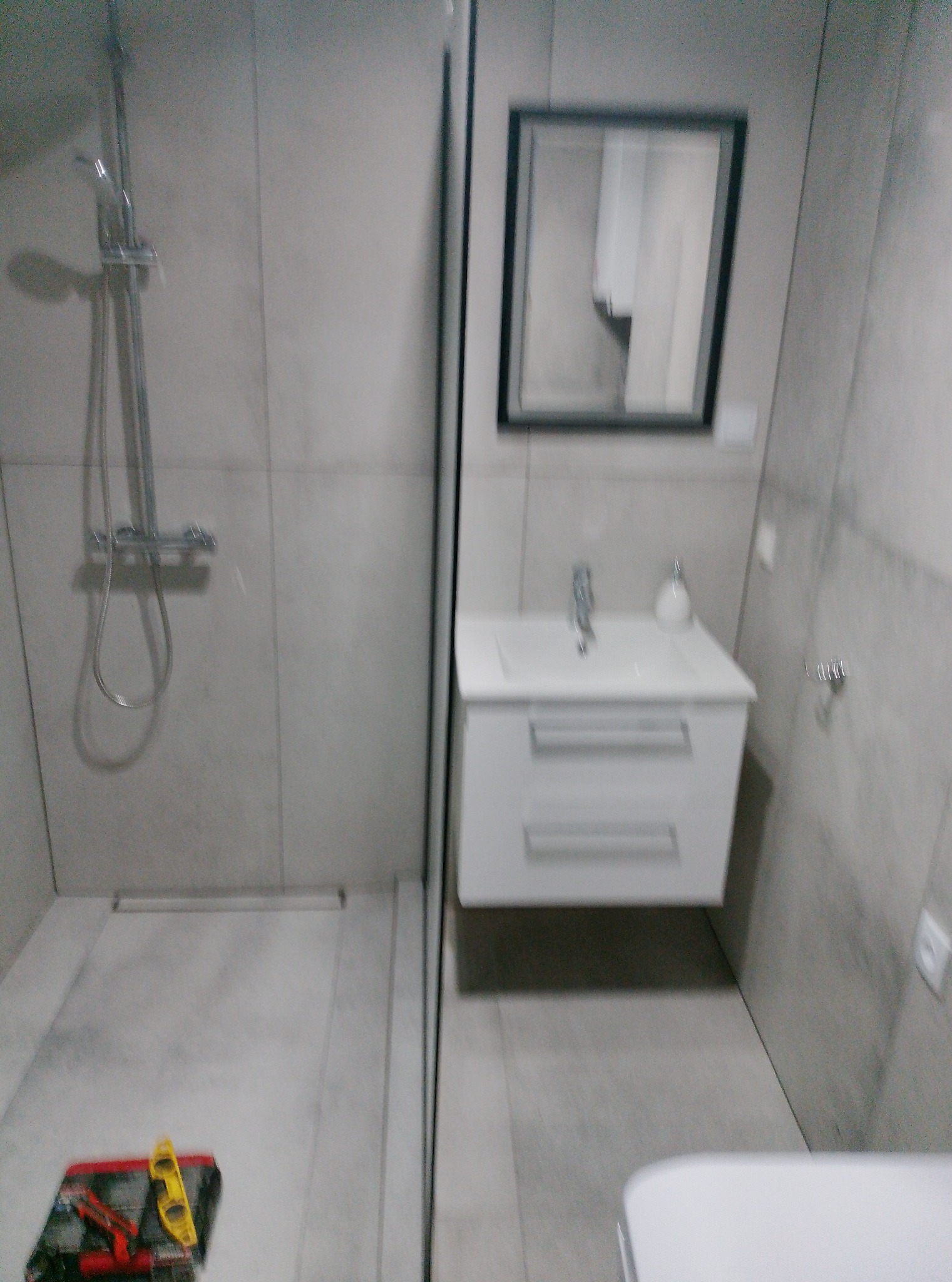 Wykończona łazienka w szarościach: kabina prysznicowa ze szklaną ścianką, biała szafka z umywalką i lustro w czarnej ramie. Na podłodze leżą narzędzia.