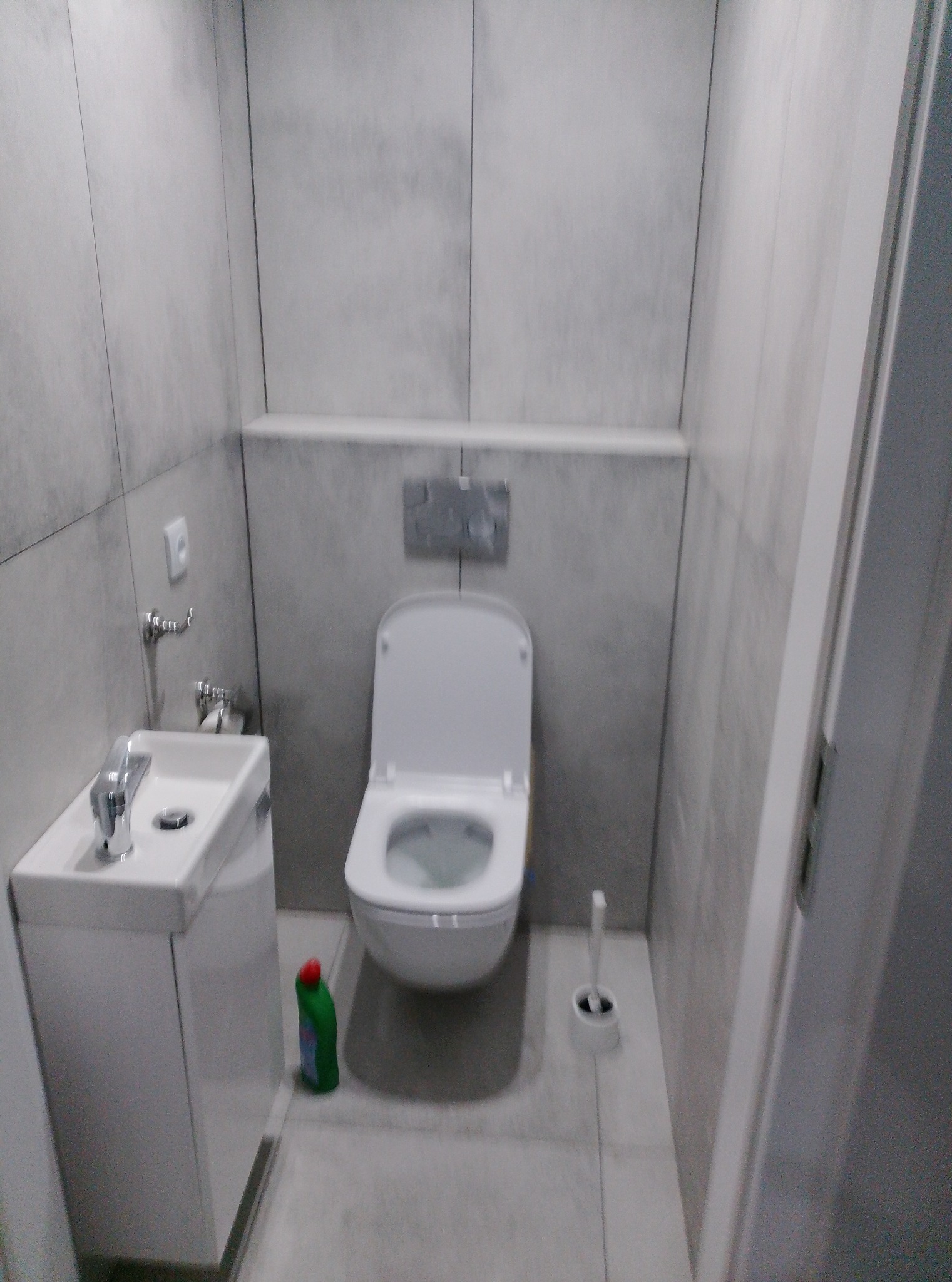 Nowoczesna, minimalistyczna łazienka w odcieniach szarości z wiszącą toaletą, umywalką z szafką i akcesoriami. Widoczne płytki imitujące beton i butelka środka czystości.