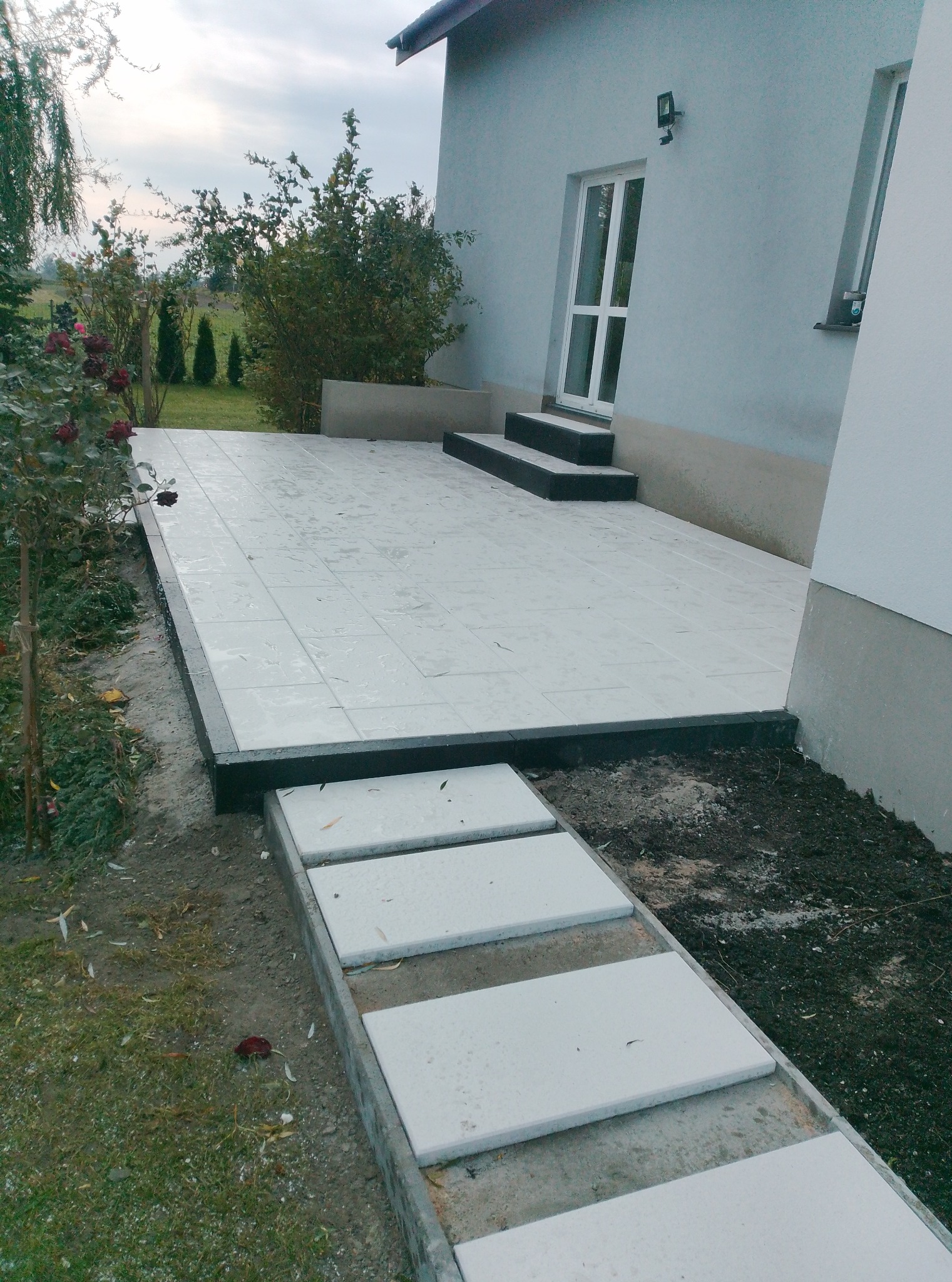 Wykończony taras z jasnych płyt, z czarną obwódką, prowadzący do drzwi wejściowych. Dodatkowo ułożony chodnik z płyt betonowych. Widok na ogród i elewację budynku.