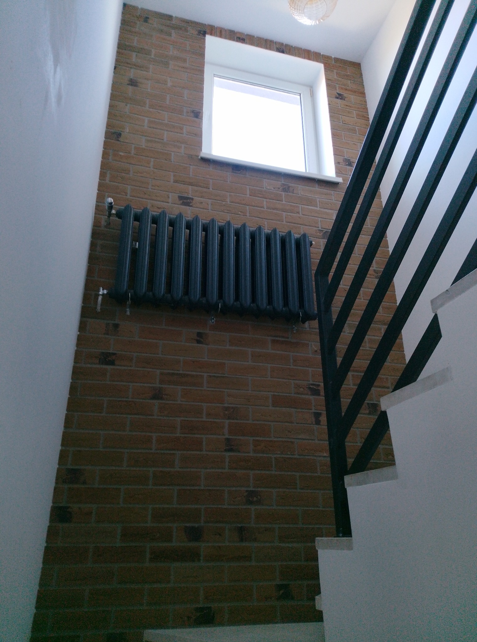 Ceglana ściana ze stalowym grzejnikiem i oknem, widok z dołu. Fragment schodów z czarną balustradą po prawej. Oświetlenie sufitowe widoczne w górnej części kadru.