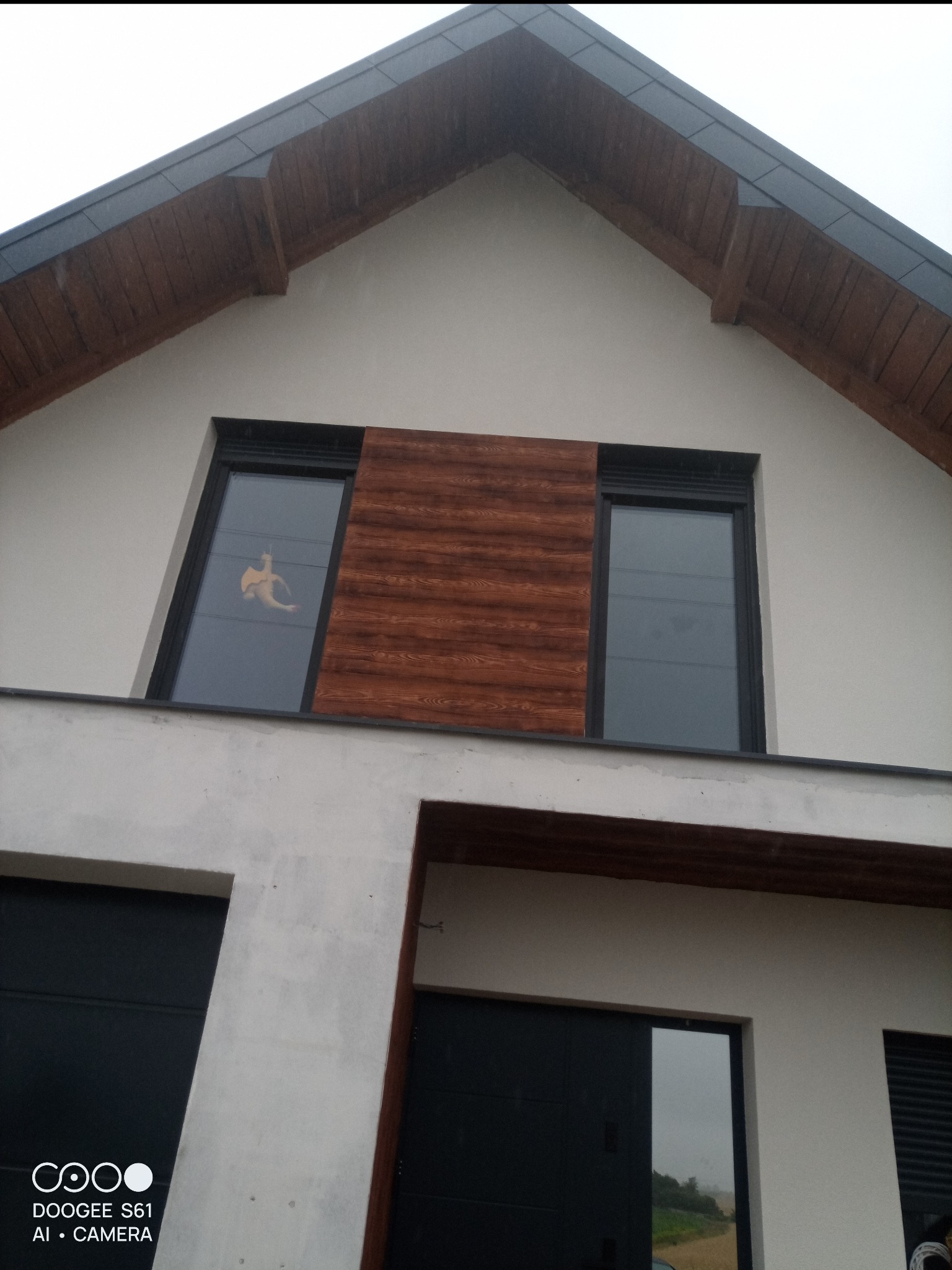 Elewacja domu z oknami i drewnianą okładziną. Widoczne czarne bramy garażowe i drewniane wykończenie dachu. W oknie naklejka.