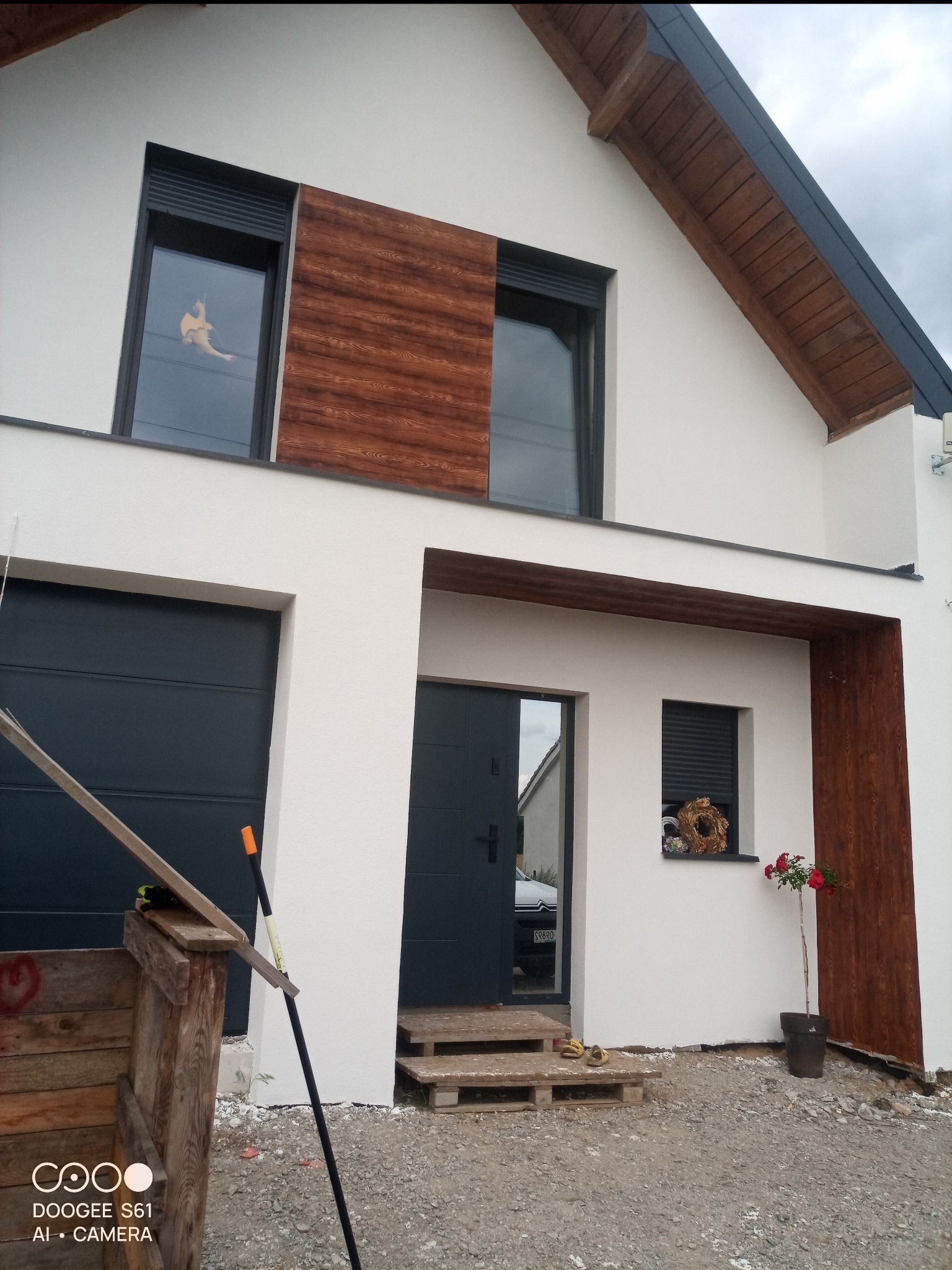 Nowoczesny dom z białą elewacją, drewnianymi akcentami i ciemnymi oknami. Widoczny fragment garażu, wejście i dekoracje okienne. Uwagę przykuwa świeżo posadzona róża.