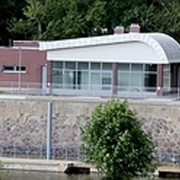 Nowoczesny budynek z przeszklonymi ścianami i zaokrąglonym dachem, widoczny z perspektywy rzeki, otoczony zielenią. Minimalistyczna architektura w miejskim krajobrazie.