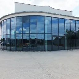 Nowoczesny budynek z fasadą ze szkła refleksyjnego odbijającą błękitne niebo i chmury, otoczony brukowanym placem i niską zielenią.