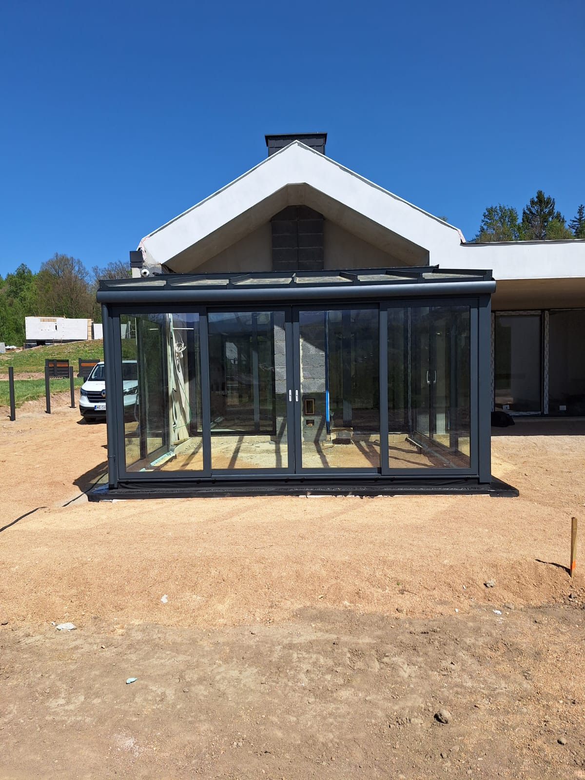 Nowoczesna, przeszklona oranżeria z przesuwnymi drzwiami w grafitowej ramie, dobudowana do budynku z białym dachem, na tle błękitnego nieba i zieleni.