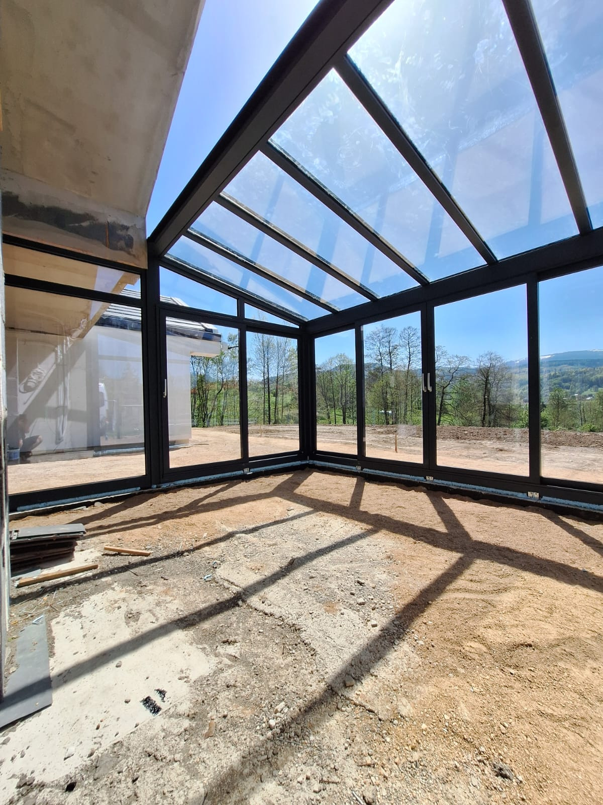 Nowoczesna, przeszklona weranda z czarnymi ramami, widok na zieleń za oknami. Wnętrze w stanie surowym, przygotowane do dalszych prac wykończeniowych.