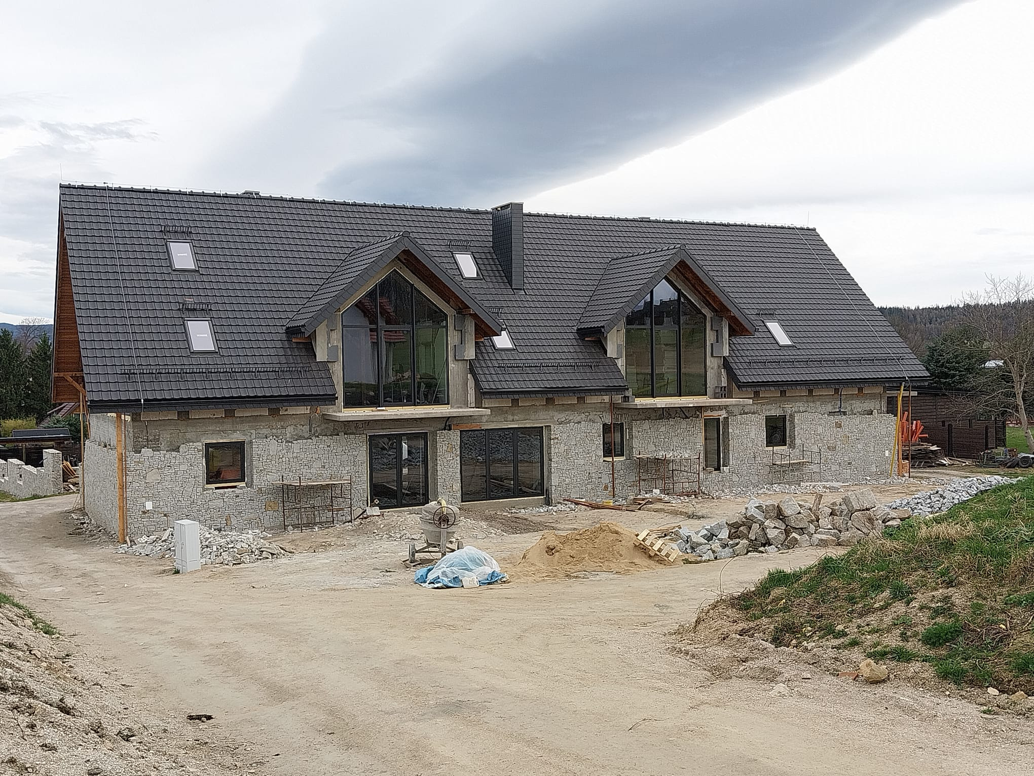 Surowy stan domu jednorodzinnego z szarą dachówką i dużymi oknami, w trakcie prac wykończeniowych elewacji. Widoczne rusztowania i materiały budowlane.