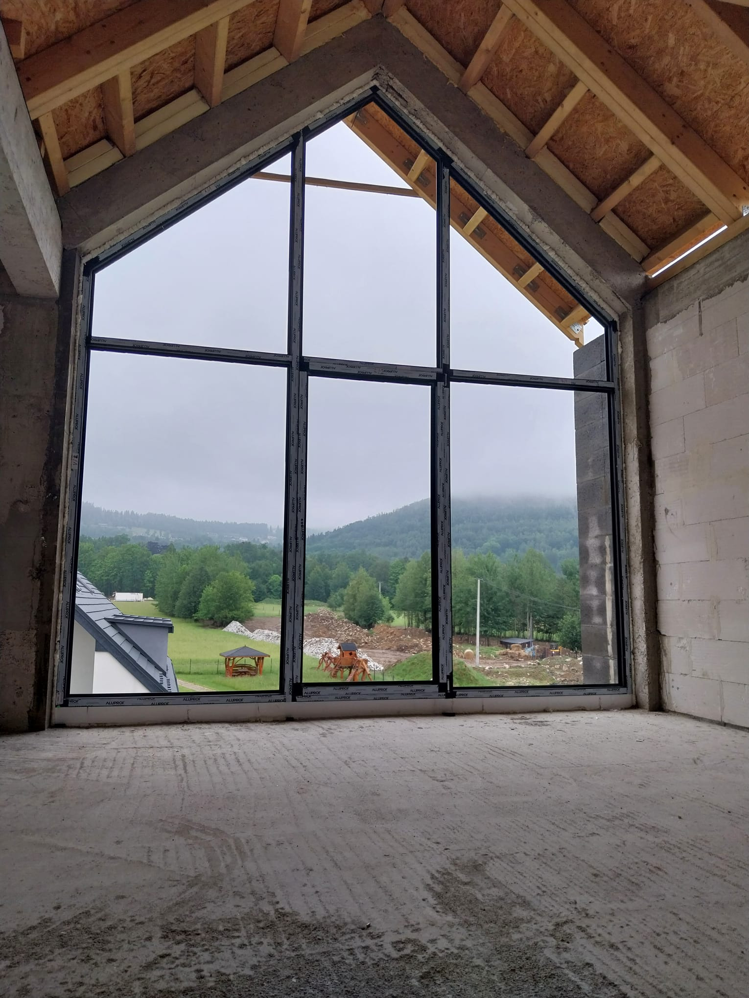 Wysokie okno z czarnymi ramami w surowym wnętrzu domu w budowie. Widok na zalesione wzgórza i zabudowania w oddali. Rama okna z widocznym napisem 'ALUPROF'.