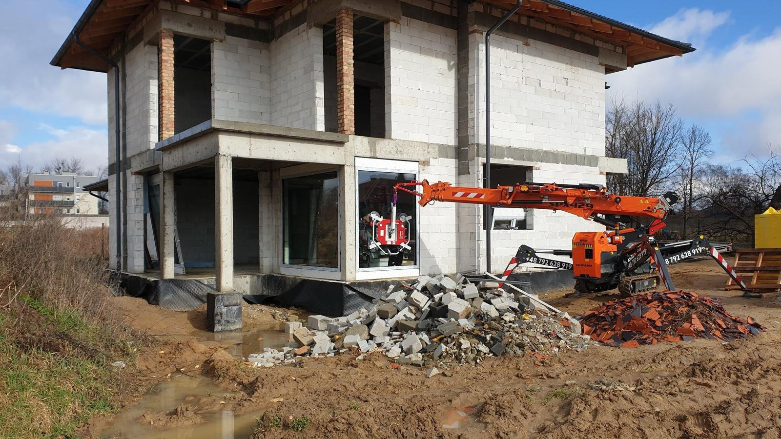 Montaż okna w surowym budynku z użyciem specjalistycznego dźwigu. Widoczne gruzy i błoto na pierwszym planie. Budynek z białego bloczka.