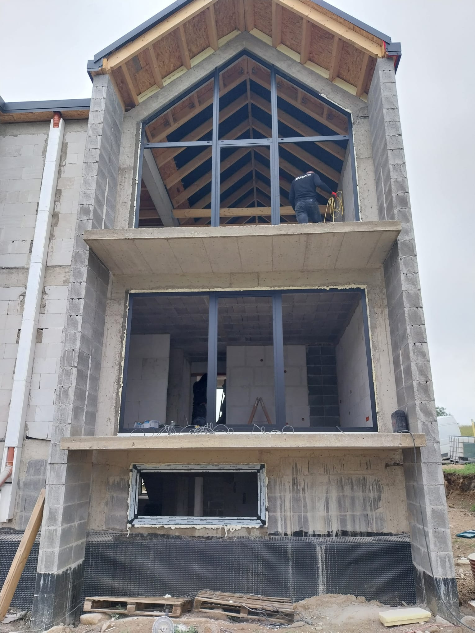 Montaż stolarki okiennej w surowym budynku. Widoczne trzy kondygnacje z oknami o czarnych ramach. Na górnej kondygnacji pracownik z kablem.