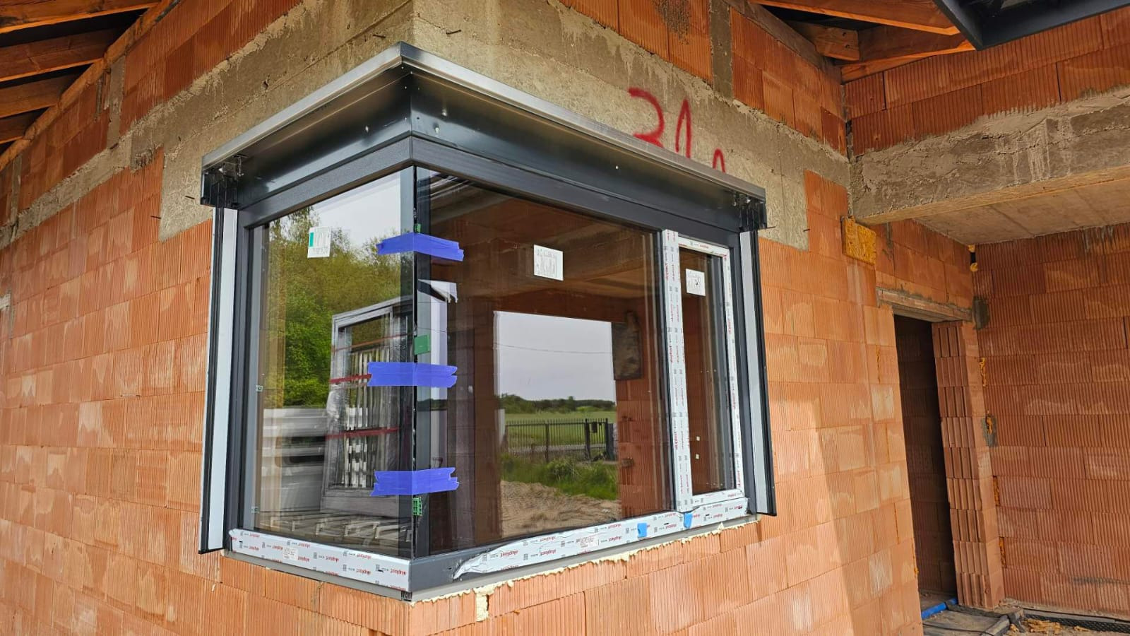 Narożne okno PCV z czarną ramą, zamontowane w surowym murze z cegły. Widoczne taśmy zabezpieczające na szybach i odblask drugiego okna wewnątrz budynku.