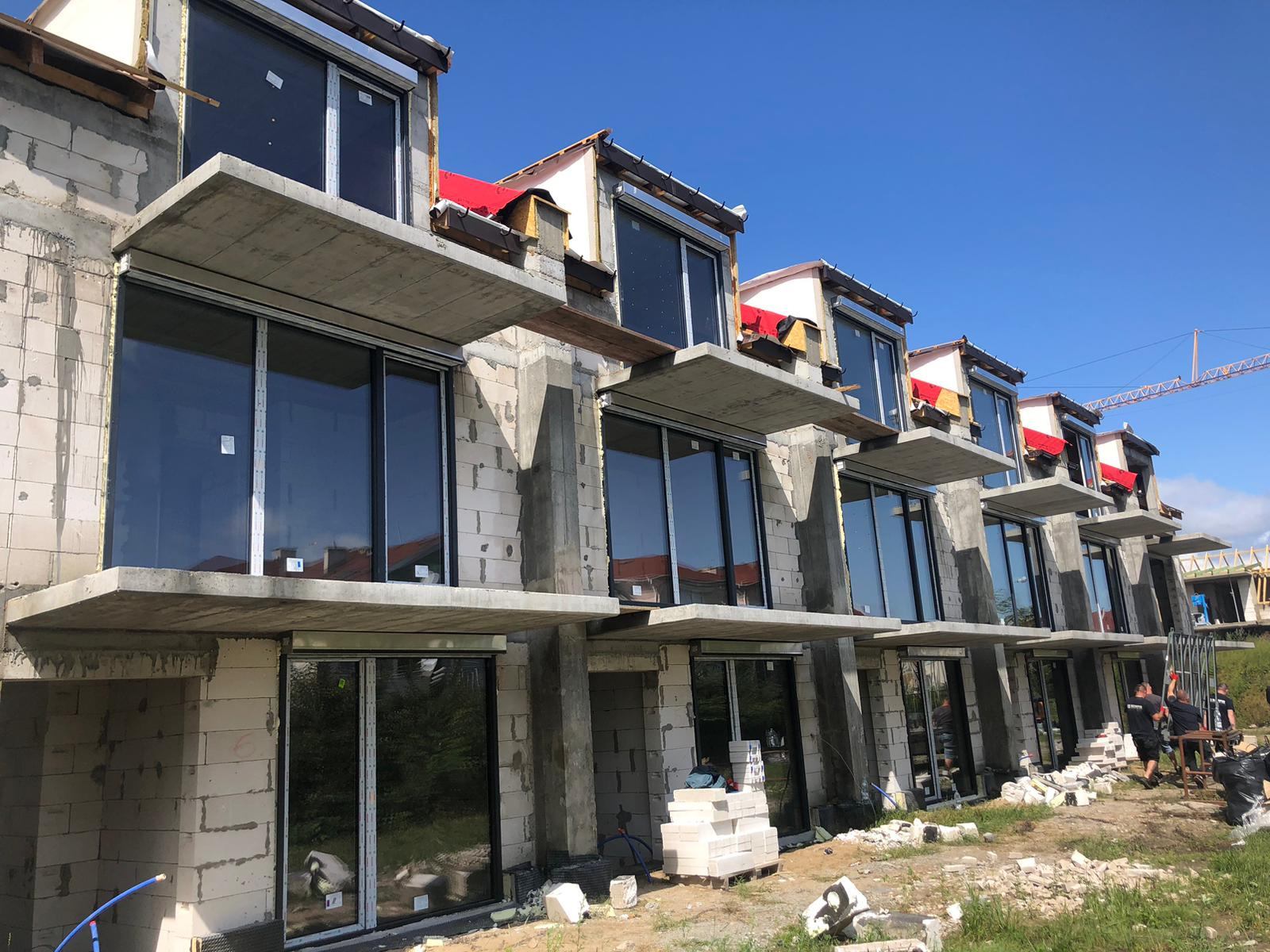 Rząd domów w trakcie budowy z zamontowanymi oknami i drzwiami przesuwnymi, widok z dołu, błękitne niebo w tle, na pierwszym planie porozrzucane materiały budowlane.