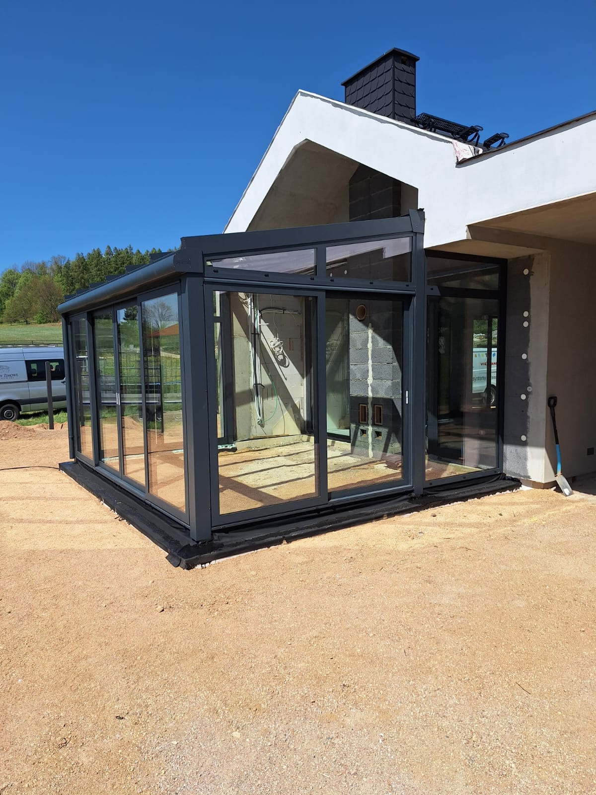 Szklana zabudowa tarasu z przesuwnymi drzwiami w grafitowej ramie, widok z zewnątrz na tle budynku w trakcie wykończenia i błękitnego nieba.