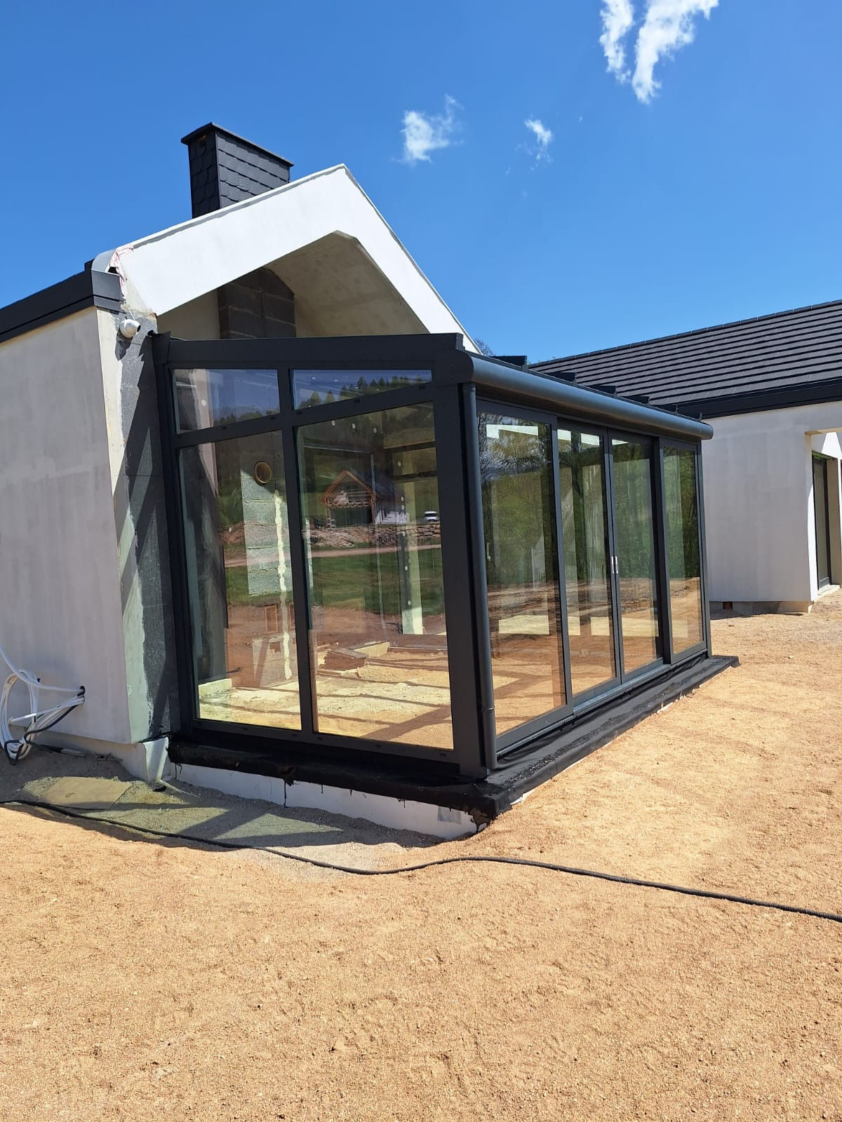 Nowoczesna, przeszklona zabudowa tarasowa z czarnymi ramami, przylegająca do budynku z jasną elewacją i ciemnym dachem, w słoneczny dzień. Widoczne przesuwne segmenty.